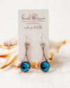 A pair of silver-toned drop earrings with round blue marbled stones, displayed on a white card labeled beach blossom studio, set against a white background with a seashell prop.