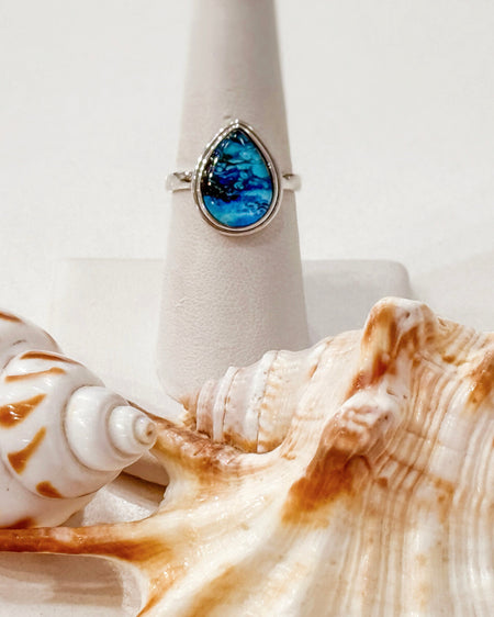 A silver ring with a teardrop-shaped blue marbled stone, displayed on a white cone stand with seashells in the foreground.